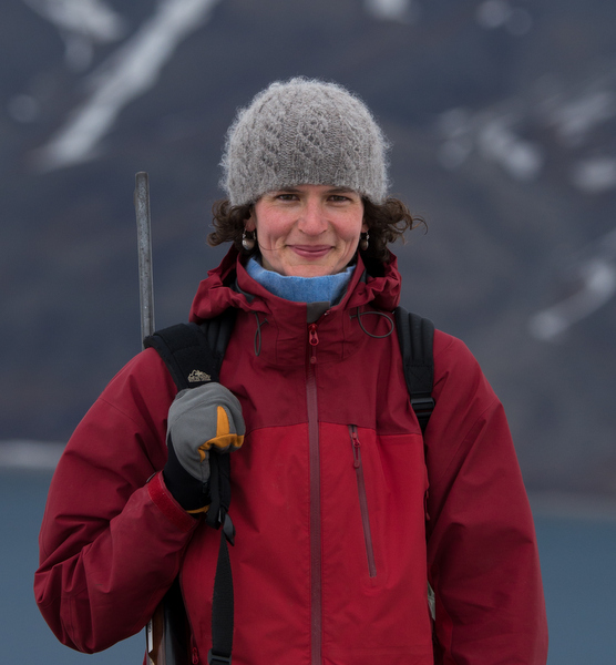 Jennifer Kingsley in her toque and winter jacket with a gun slung over her shoulder and a mountain and lake in the background.