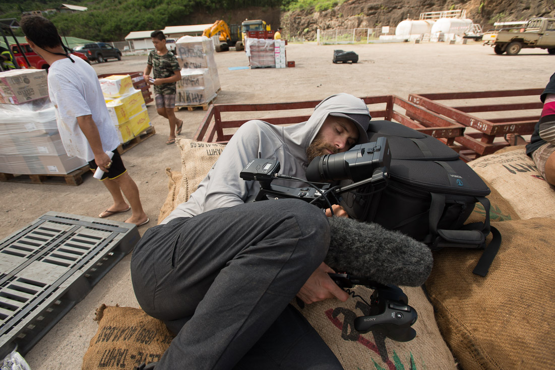 A cameraman lies on the ground in a construction site to get his shot while two people stand a few feet away in the background.