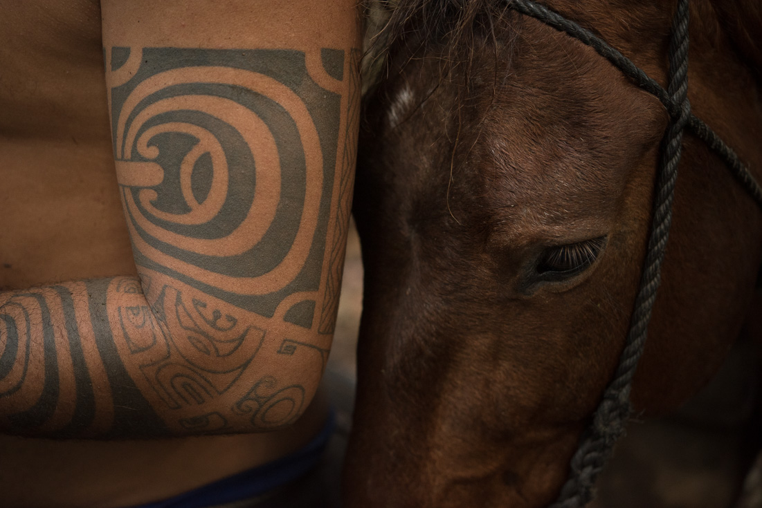 A man stands next to a horse and shows off the traditional tattoos on his upper arm.